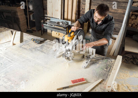 A young male carpenter builder saws a modern circular saw a wooden board in the workshop room Stock Photo