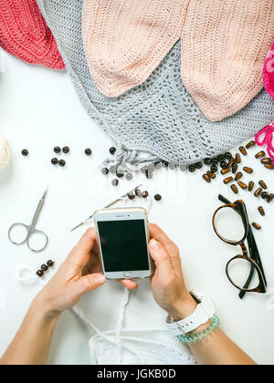 A close-up on top is a table for creativity with knitted things, threads, beads, glasses, ribbons. Young woman holds a phone in hands Stock Photo