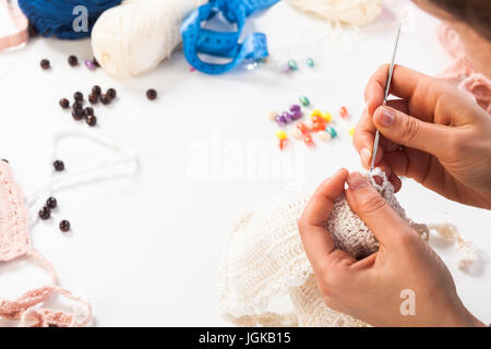 A close-up on top is a table for creativity with knitted things, threads, beads, glasses, ribbons. Young woman knits a crochet of white thread scarf Stock Photo