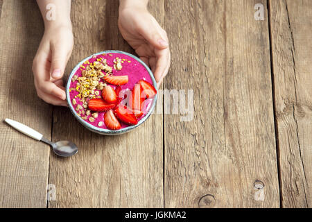 Eating healthy breakfast bowl. Acai smoothie, granola, seeds, fresh strawberries in ceramic bowl in female (child) hands over wooden background. Clean Stock Photo