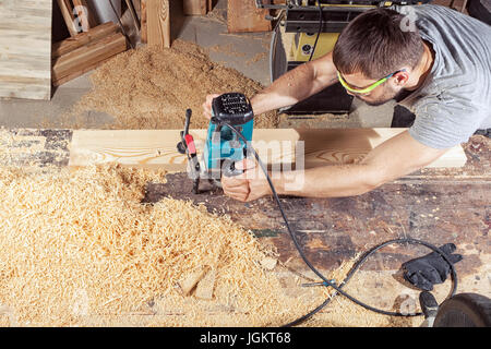 A young brunette man in safety glasses and a gray T-shirt is working a wooden board with a milling machine in the workshop, around a lot of wooden saw Stock Photo