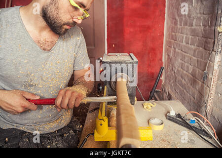 A young brunette man builder wearing green protective goggles, a gray T-shirt and with a beard working on a wooden workpiece on a lathe in the worksho Stock Photo