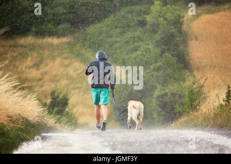 Young man running with his dog (labrador retriever) in heavy rain on the rural road. Stock Photo