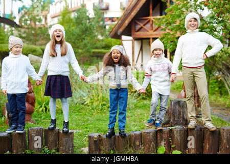 Happy children of various ages having fun playing outdoors in green countryside, standing in row on wooden posts holding hands dressed in similar whit Stock Photo