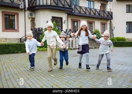 Group of happy children playing outdoors and running towards camera holding hands, all wearing similar knit clothes on warm autumn day Stock Photo