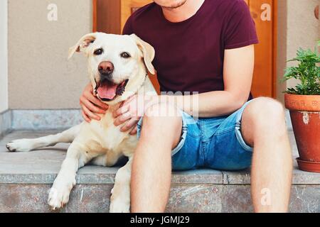 Happy labrador retriever. Young man resting with old dog on the stairs in front of the house. Stock Photo