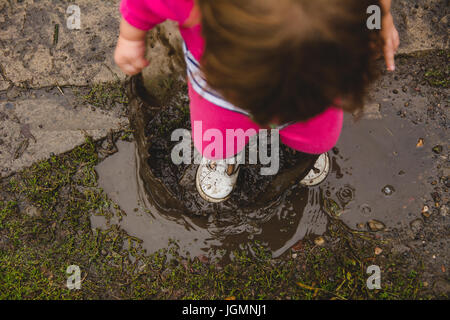 A 2-year old girl, looking down at ground and mud puddle, splashes in a mud puddle, making a mess of her clothes and shoes Stock Photo