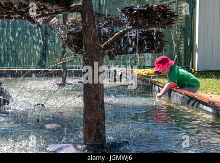 Young child wearing red sunhat leaning into water fountain water at RHS Hampton Court Flower Show, London, England, UK Stock Photo