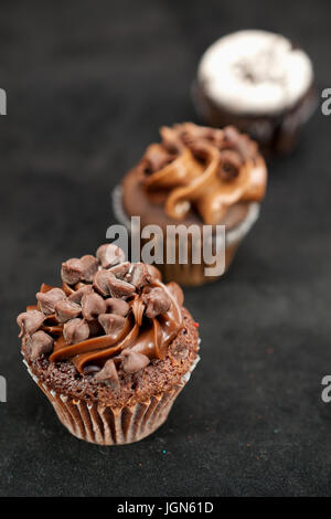 Three chocolate cupcakes garnished with chocolate chips and whipped cream frosting in row across the frame, on black background Stock Photo