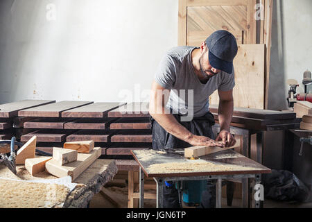 A young brunette man with a beard in a cap and T-shirt by profession carpenter builder processes the edges of a wooden product using a milling machine Stock Photo