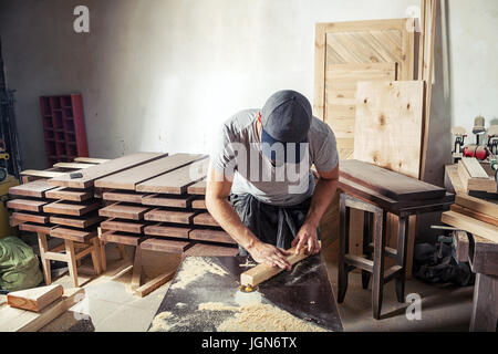 A young brunette man with a beard in a cap and T-shirt by profession carpenter builder processes the edges of a wooden product using a milling machine Stock Photo