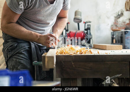 A close-up as a young man holds a black jack plane in his hands and processes a wooden block, in the background a lot of wooden boards and equipment Stock Photo