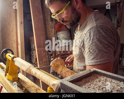 A young man with dark hair and goggles holds a stalker in his hands and processes a wooden product on a lathe in the workshop Stock Photo