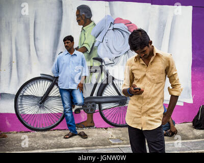 July 9, 2017 - Singapore, Singapore - Guest worker from the Indian sub-continent in front of a mural in Singapore. There are hundreds of thousands of guest workers from the Indian sub-continent in Singapore. Most work 5 Â½ to six days per week. On Sundays, the normal day off, they come into Singapore's ''Little India'' neighborhood to eat, drink, send money home, go to doctors and dentists and socialize. Most of the workers live in dormitory style housing far from central Singapore and Sunday is the only day they have away from their job sites. Most work in blue collar fields, like constructio Stock Photo