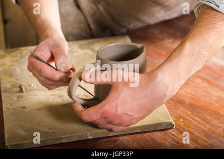 pottery, stoneware, ceramics art concept - closeup on craftsman hands connecting handle to cup with tool, master fingers works with pieces of fireclay Stock Photo