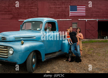 American Farm family with vintage blue truck in front of red barn with American flag, dog at feet, red headed toddler Stock Photo