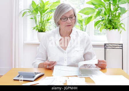 Senior german woman at home with a calculator and lots of bills and receipts, filling out the printed forms of the annual tax declaration Stock Photo