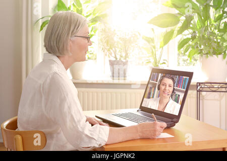 Senior woman in her sunny living room in front of a laptop making notes during watching an online advice video by a female therapist Stock Photo