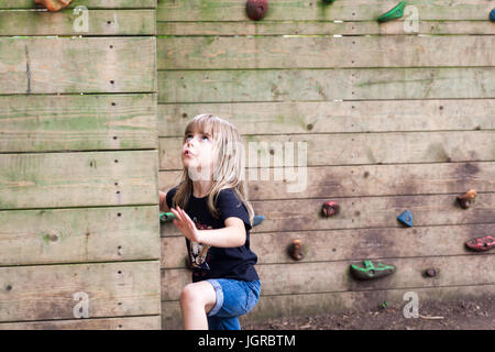 Little girl starting to climb up a wooden climbing wall. Cute child with physical active lifestyle outdoors Stock Photo