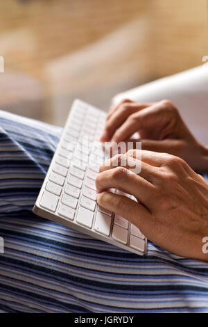 closeup of a young caucasian man in pajamas sitting in a comfortable armchair typing in a computer keyboard Stock Photo