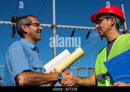 Smiling workers with blueprints and clipboard in meeting at electrical substation. Stock Photo