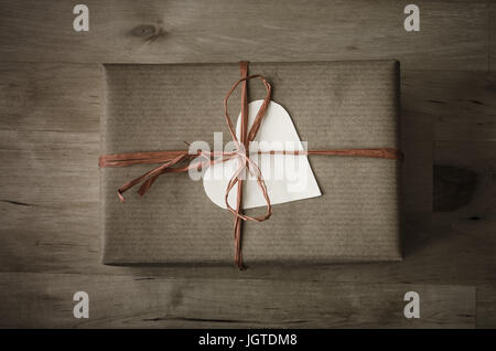 Overhead shot of a gift box with a heart shaped blank label, wrapped in brown paper with a raffia ribbon tied to a bow on wooden planked table.  Vinta Stock Photo