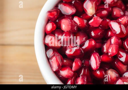 Overhead macro shot of a white bowl filled with red pomegranate seeds on a wooden table. Stock Photo