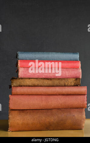 Close shot of a pile of old, shabby, well used text books stacked in a pile on a desk in front of a black chalkboard.  Copy space above. Stock Photo