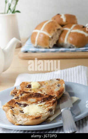 Spring or Easter breakfast table with a serving of toasted, sliced and buttered hot cross buns on plate with knife and napkin;.  Visible in the backgr Stock Photo