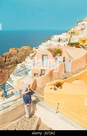 woman wearing hat, blue top and white pants standing on terrace with her back to camera and looking at beautiful houses of Oia on sunny summer day, Sa Stock Photo