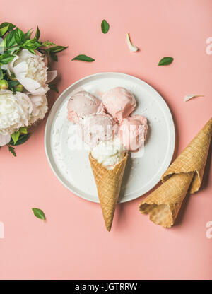 Flatlay of pastel pink strawberry and coconut ice cream scoops, sweet cones on white plate and white peonies over pastel pink background, top view, ve Stock Photo