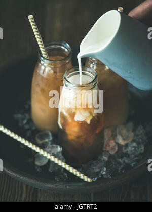 Refreshing summer drink. Cold Thai iced tea in glass bottles with milk pouring from jug on plate over dark wooden background, selective focus. Vegetar Stock Photo