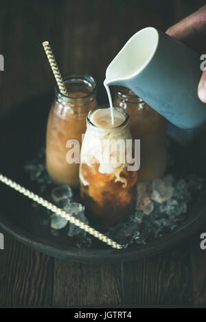 Refreshing summer drink. Cold Thai iced tea in glass bottles with milk pouring from white jug on plate over dark wooden background, selective focus. V Stock Photo