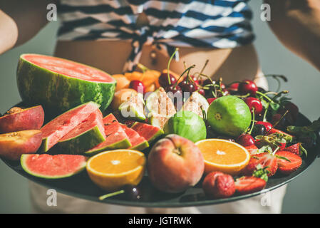 Summer healthy raw vegan clean eating breakfast in bed concept. Young girl wearing striped home shirt holding tray full of fresh seasonal fruit, selec Stock Photo