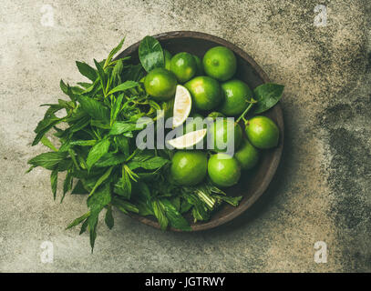 Flatlay of freshly picked organic limes and mint leaves for making cocktail or lemonade in wooden plate over grey concrete stone background, top view Stock Photo