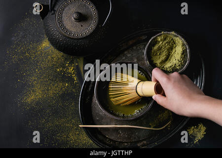 Green tea matcha powder and hot drink in black bowls standing with iron teapot, bamboo traditional tools spoon and whisk in hand in vintage tray over Stock Photo