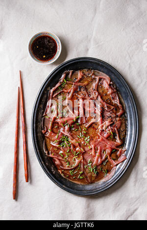Asian style Beef carpaccio with soy sauce, chive onion, black sesame, served on vintage metal tray with bowl of sauce and chopsticks over white textil Stock Photo