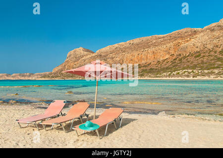 three pink sunbeds with blue hat on and umbrella on beach at Balos Bay on island of Crete in evening sun, Greece Stock Photo