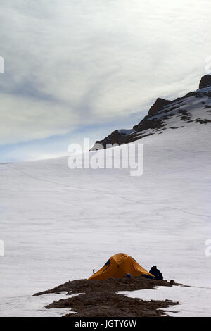 Hiker sitting near orange camping tent in evening snow mountains on overnight stay. Turkey, Central Taurus Mountains, Aladaglar (Anti-Taurus), plateau Stock Photo