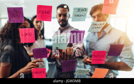 Business investors discussing work looking at the information on sticky notes stuck in office. Colleagues standing in front of post-it notes stuck on  Stock Photo