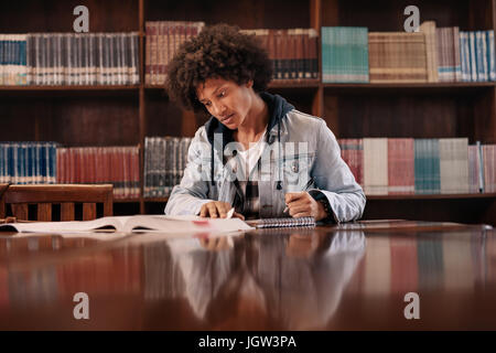 Young afro american man sitting at table with books for finding information in library. Young student making notes from books for study in library. Stock Photo