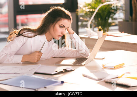 Tired young businesswoman holding pencil and looking at laptop at workplace Stock Photo