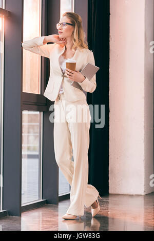 young businesswoman in suit standing in office and holding digital tablet and coffee cup Stock Photo
