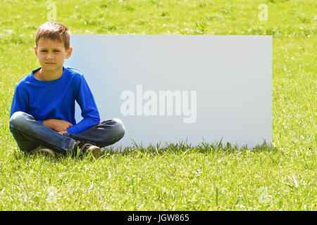 Boy sitting on the grass against blank white placard board outdoors Stock Photo