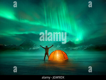 A man camping in wild northern mountains with an illuminated tent viewing a spectacular green northern lights aurora display. Photo composition. Stock Photo