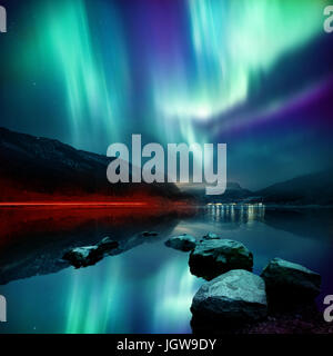A large Northern Lights (aurora borealis) display glowing over a mountain pass and reflected on a lake at night. Photo composition. Stock Photo