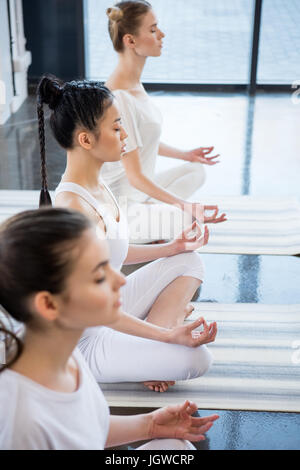 multiethnic women with eyes closed meditating in lotus pose with gyan mudra indoors Stock Photo