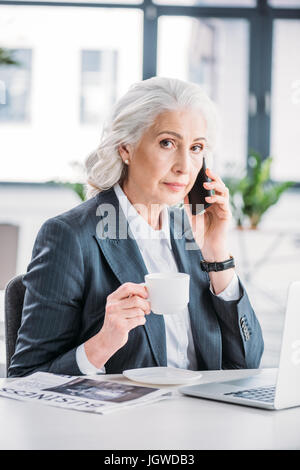 Serious senior businesswoman holding coffee cup and talking on smartphone Stock Photo