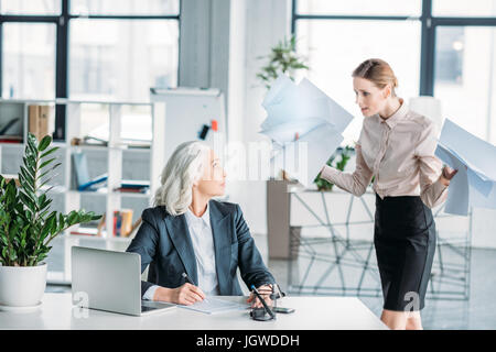 young stressed businesswoman with documents arguing at colleague sitting at workplace in office Stock Photo