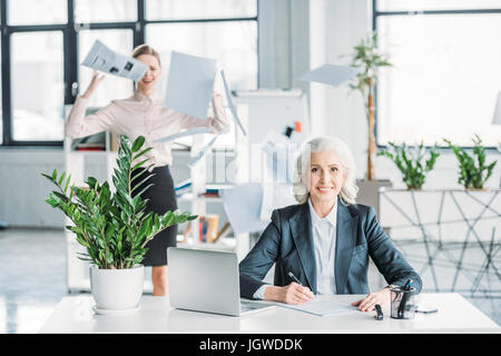 smiling businesswoman sitting at workplace in office, stressed colleague with documents behind Stock Photo